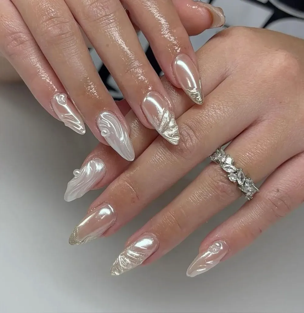 Pearl-chrome almond nails with gold-leaf marble swirls and sculpted 3D detail.