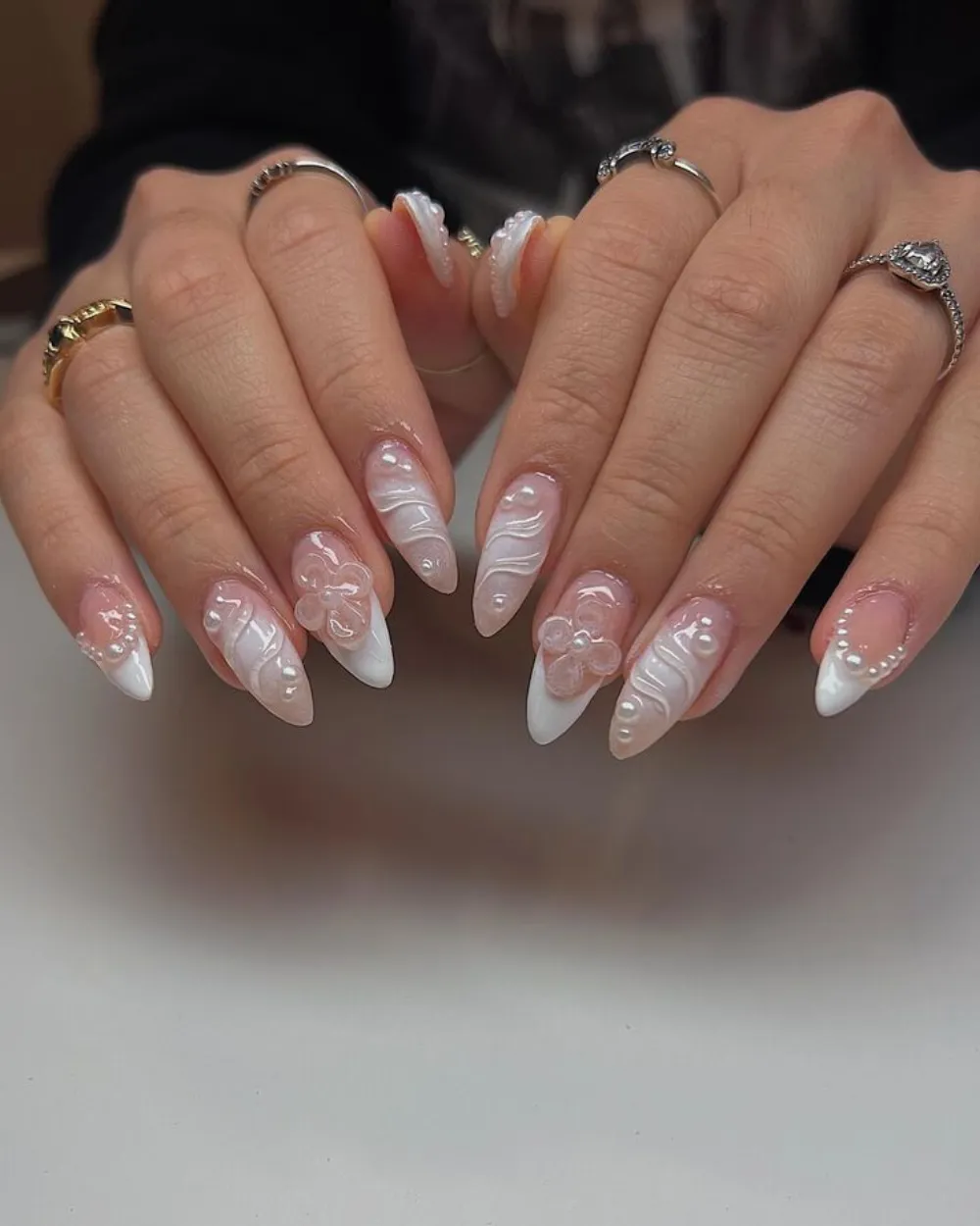 Milky white-and-nude almond nails with sculpted 3D swirls, pearls, and white floral art.
