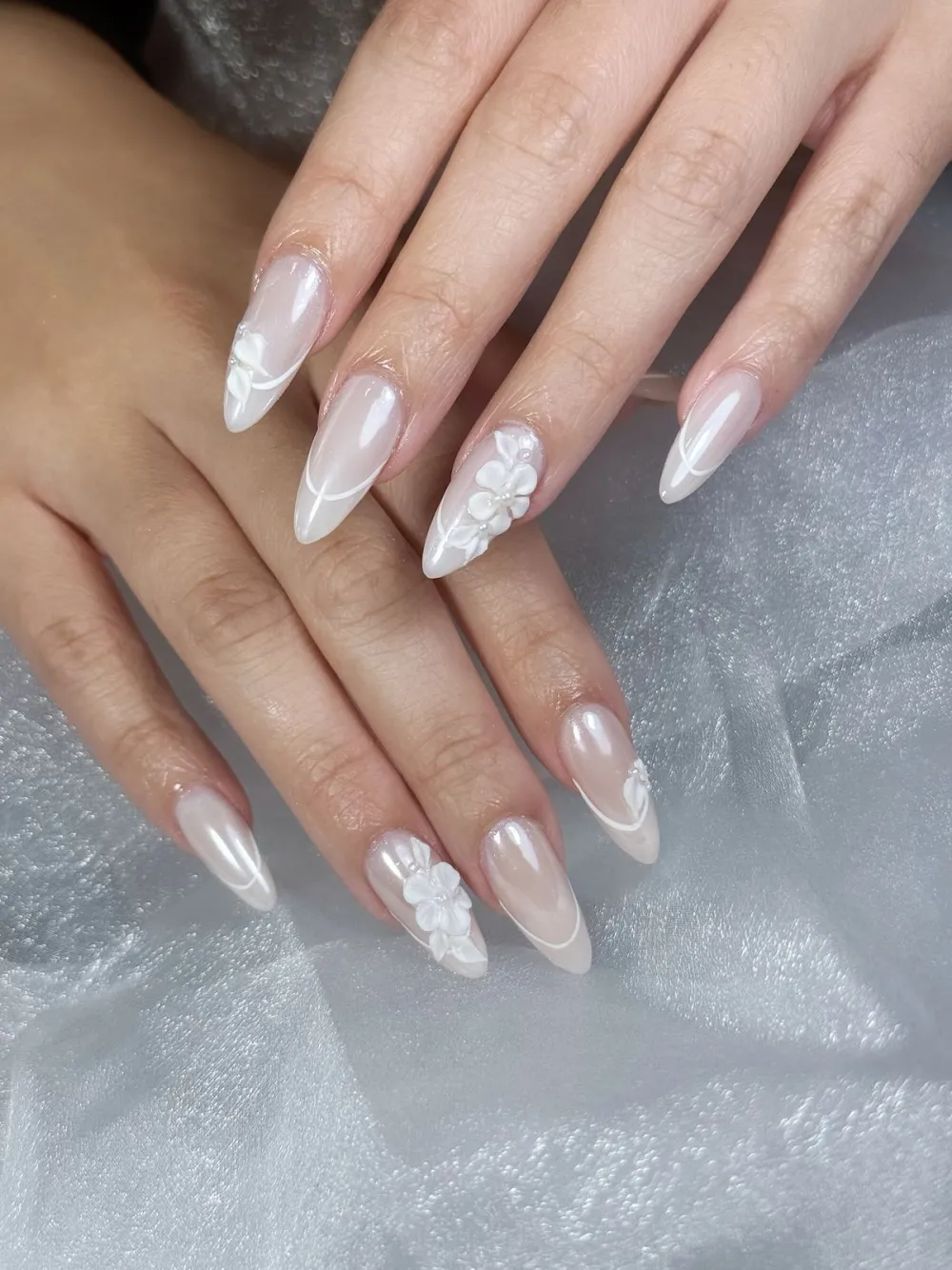 Glossy nude pearl-chrome nails with 3D white flowers on a soft tulle backdrop.