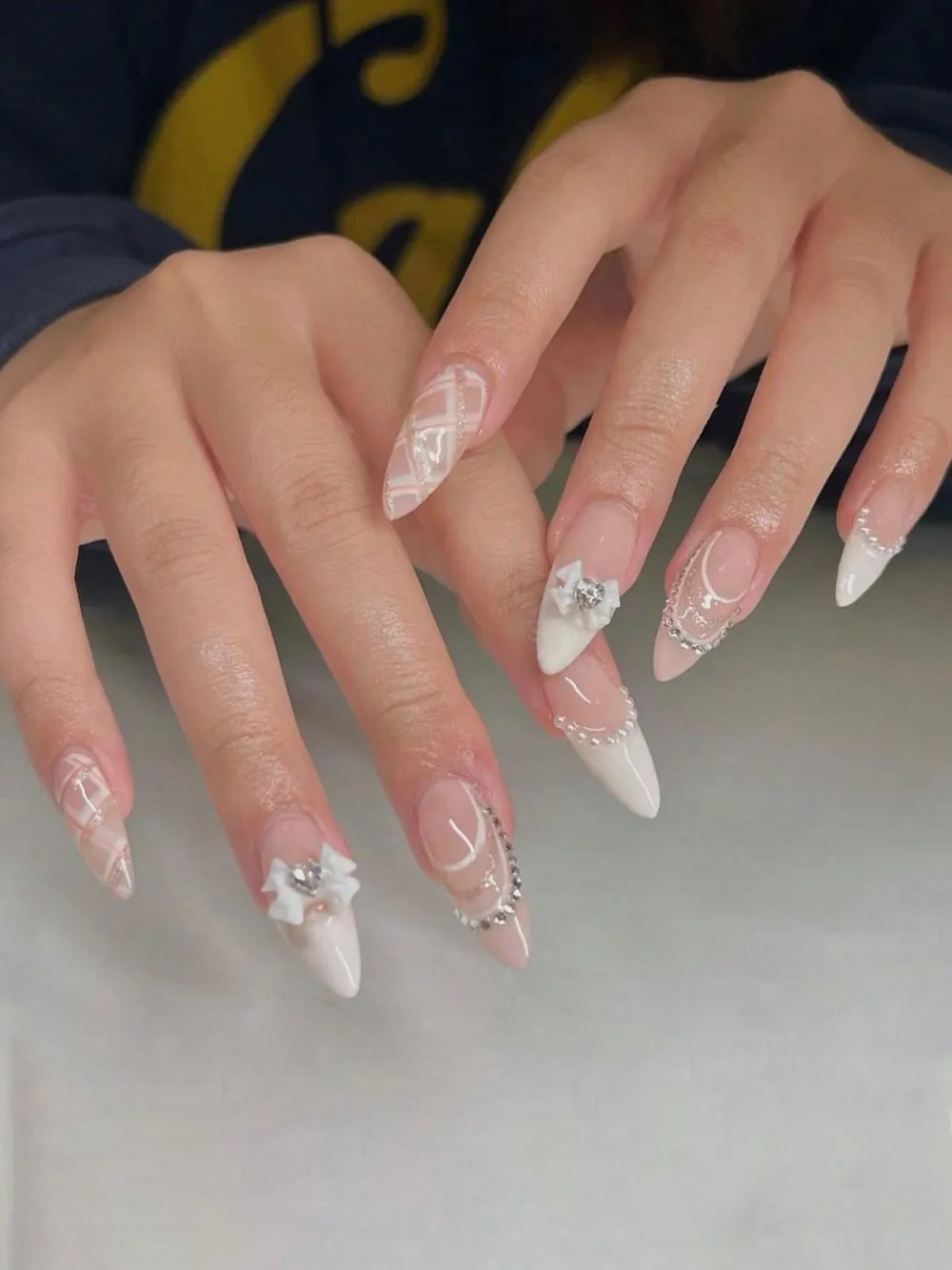Long stiletto nails in nude-and-white French style with 3D bows and rhinestone chains.